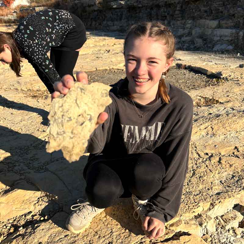 Girl holding a rock toward the camera while another girl behind her looks for rocks.