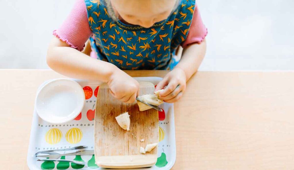 overhead view of young girl sitting at table cutting a banana on a tray.
