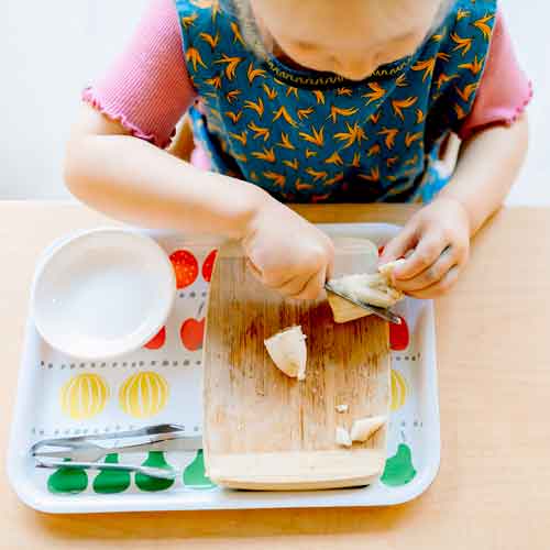 Overhead view of a toddler slicing up a banana on a tray.